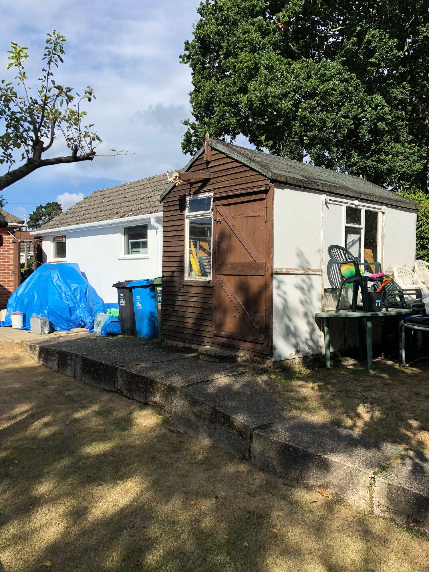 Old timber and white-rendered outbuilding beside a bungalow, with a blue tarpaulin and bins nearby